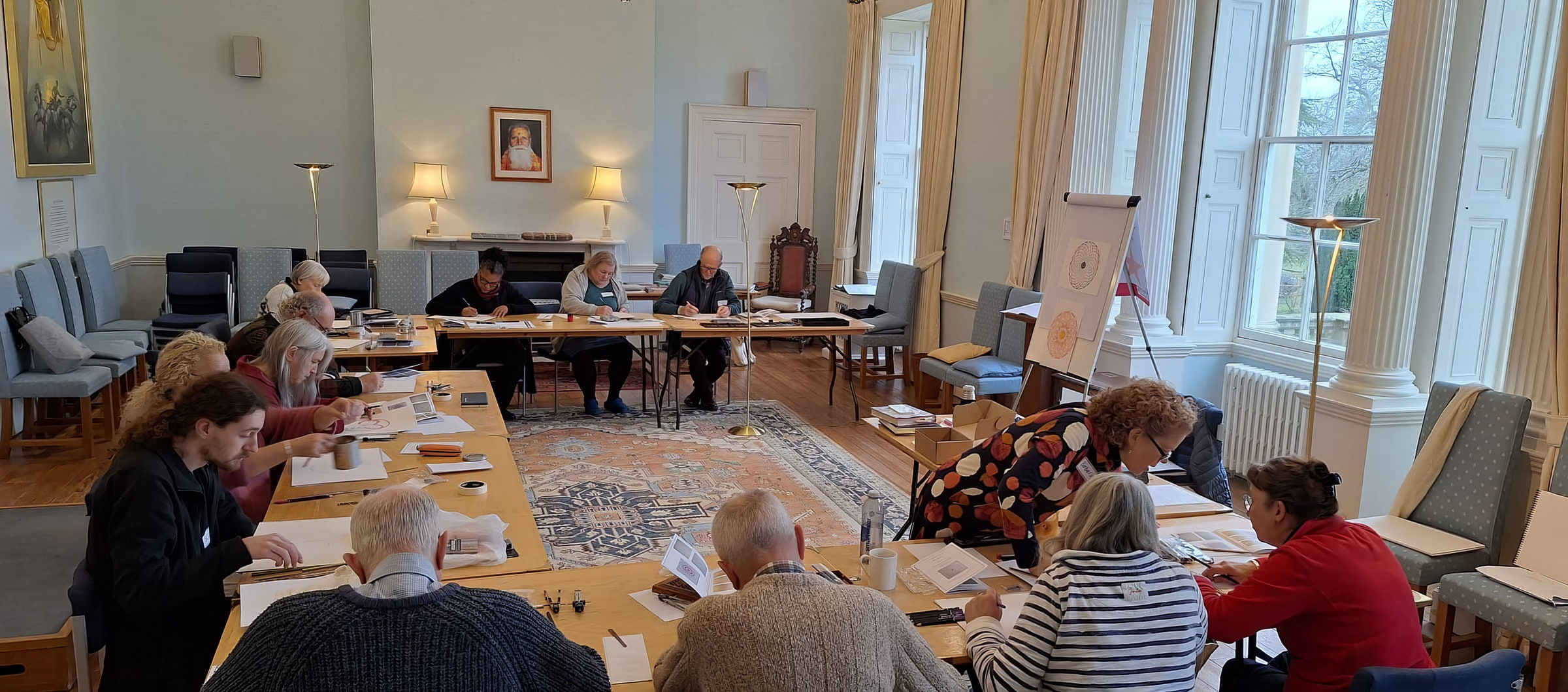 Participants drawing geometric patterns with compass and straightedge at long tables in a sunlit hall