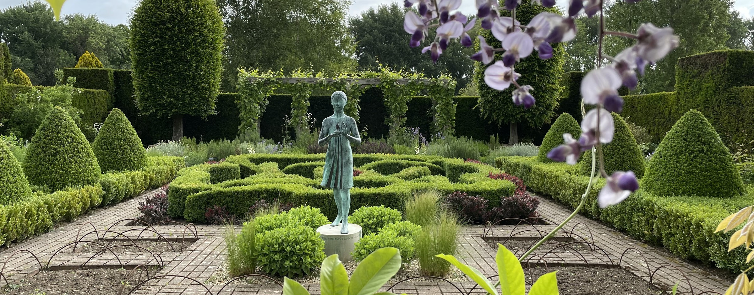 Bronze statue of a girl at the centre of a formal knot garden with conical topiary